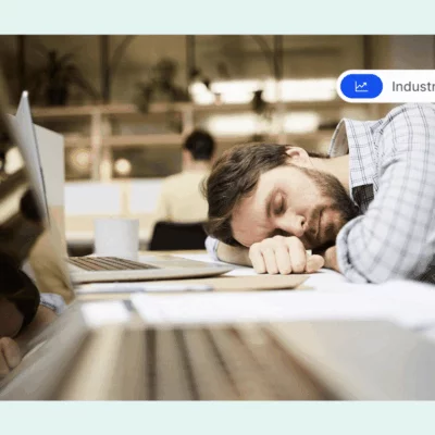 A disengaged employee sleeping at his desk in an office, with his head resting on his arm next to a laptop.