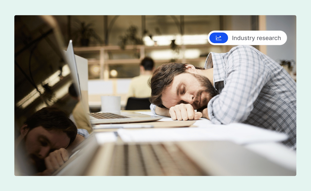 A disengaged employee sleeping at his desk in an office, with his head resting on his arm next to a laptop.