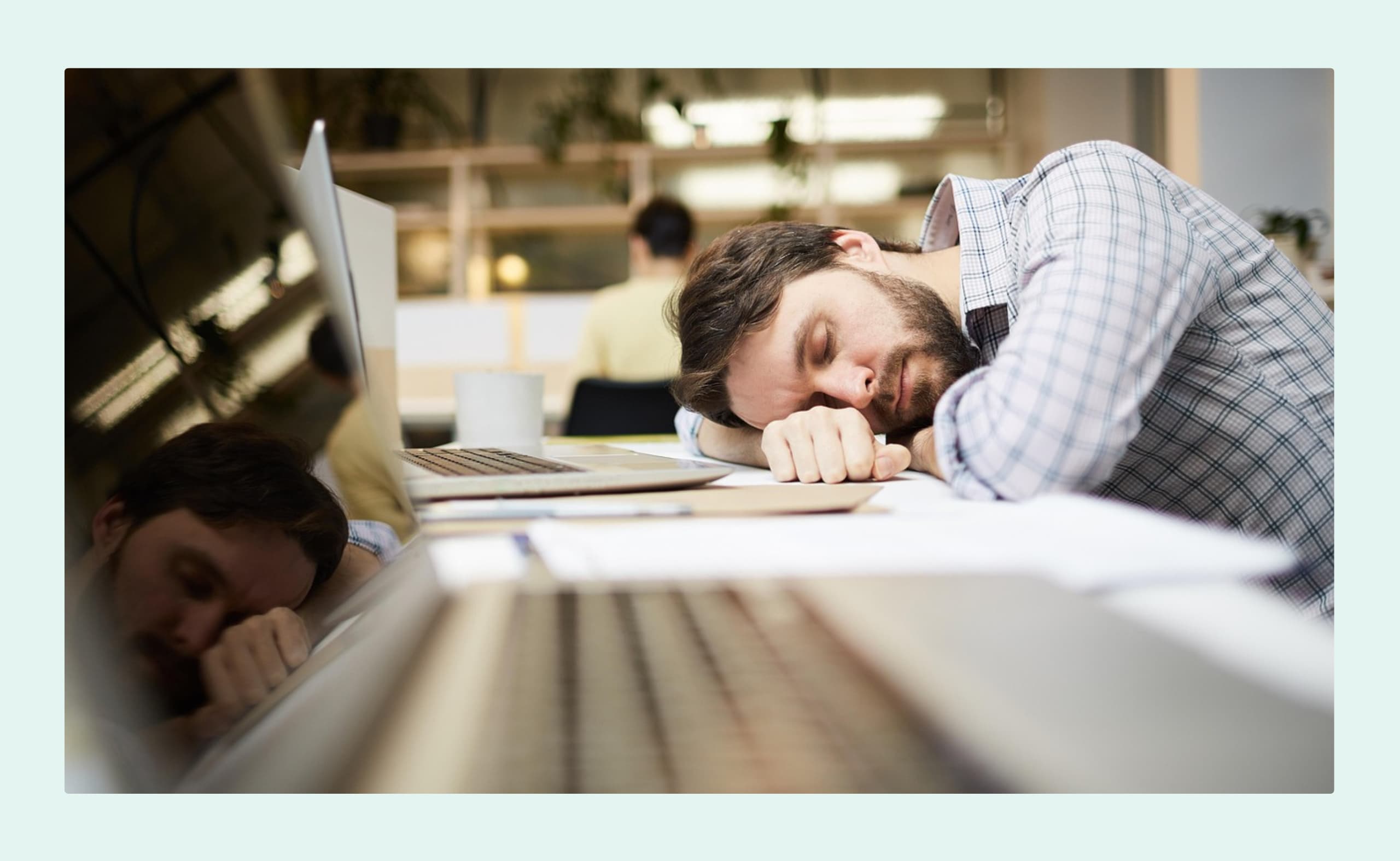 A disengaged employee sleeping at his desk in an office, with his head resting on his arm next to a laptop.