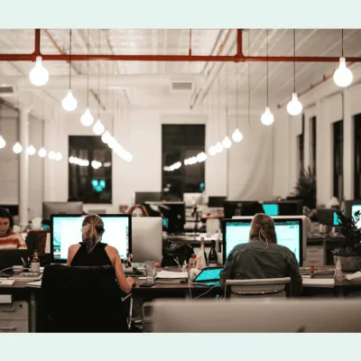 Open-plan office with people working at shared desks, surrounded by monitors and warm hanging lights in a modern coworking space.