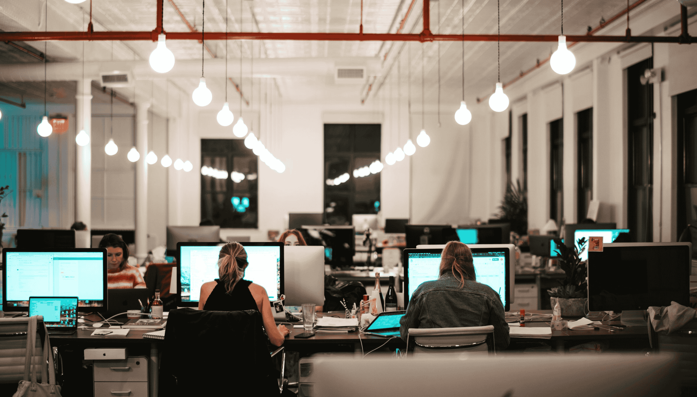 Bureau ouvert où les employés travaillent à des bureaux partagés, entourés d'écrans et de lampes suspendues chaleureuses, dans un espace de coworking moderne.