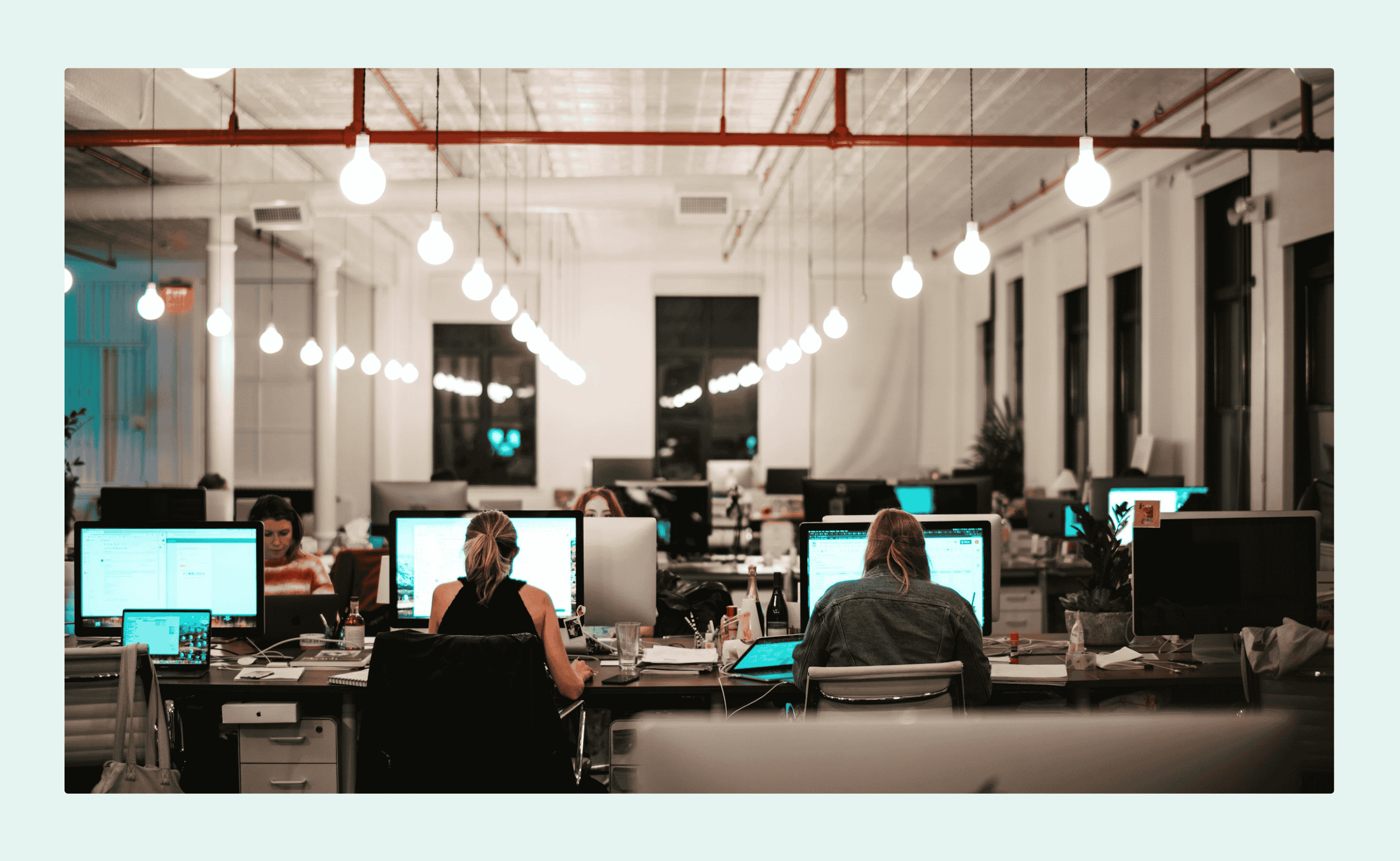 Open-plan office with people working at shared desks, surrounded by monitors and warm hanging lights in a modern coworking space.