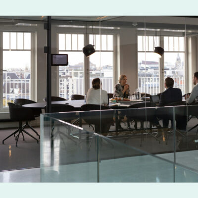 Four professionals in a glass-walled meeting room having a discussion, with city views in the background.