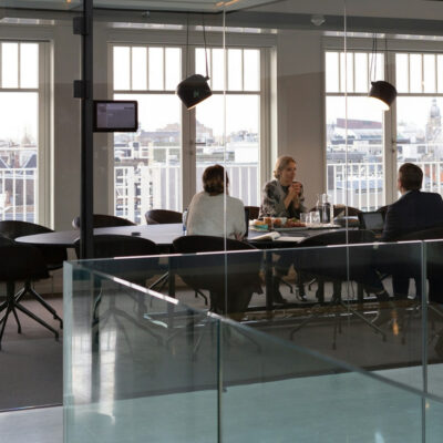 Four professionals in a glass-walled meeting room having a discussion, with city views in the background.