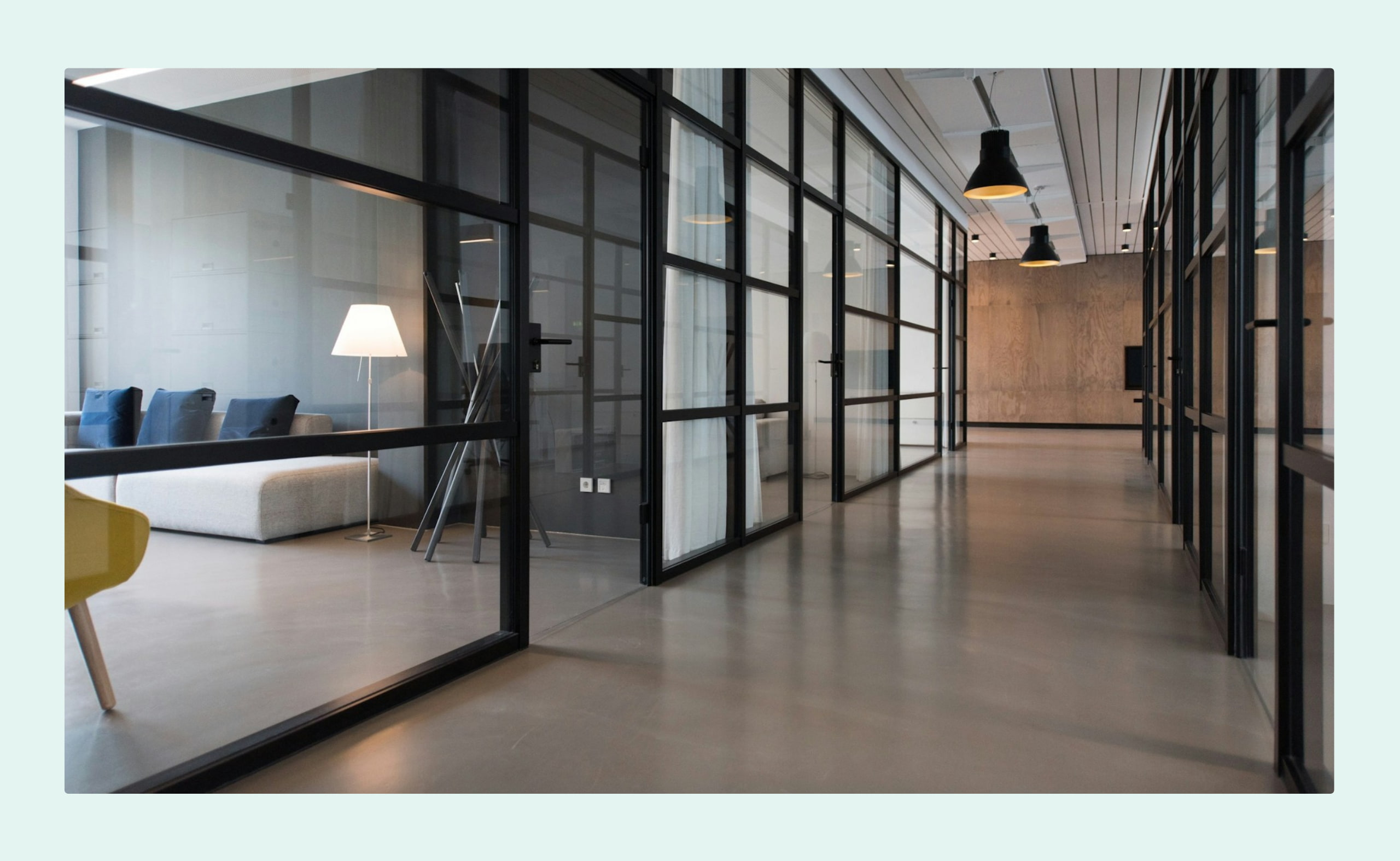 A modern office hallway with glass-walled meeting rooms, minimalist furniture, and overhead lighting.
