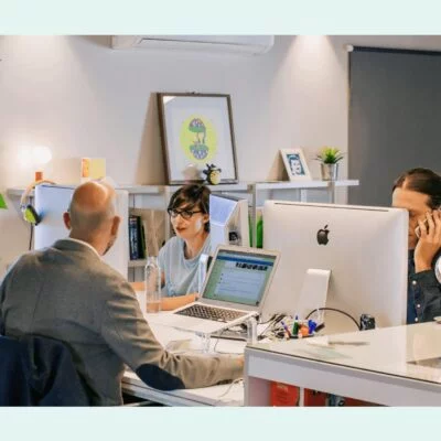Three coworkers collaborating in a bright, plant-filled modern office, with laptops and desktop computers on shared desks.