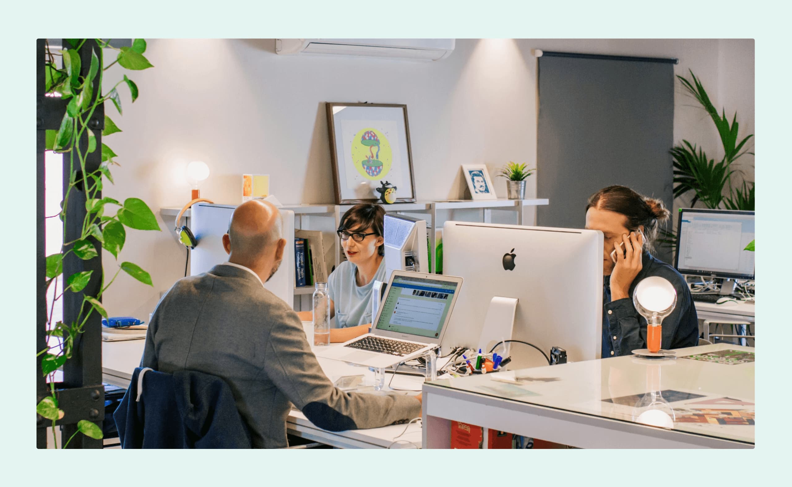 Three coworkers collaborating in a bright, plant-filled modern office, with laptops and desktop computers on shared desks.