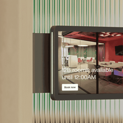 A digital room display panel showing a meeting room's availability and a “Book now” button.