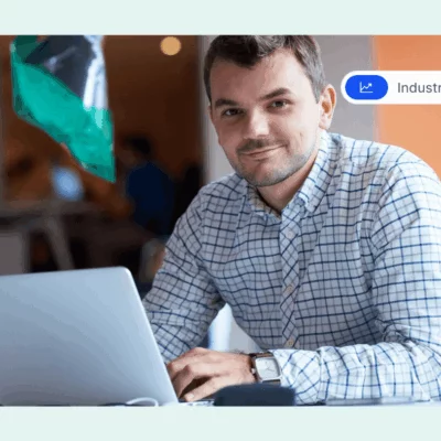 Man in a checkered shirt working on a laptop and smiling in a modern office.