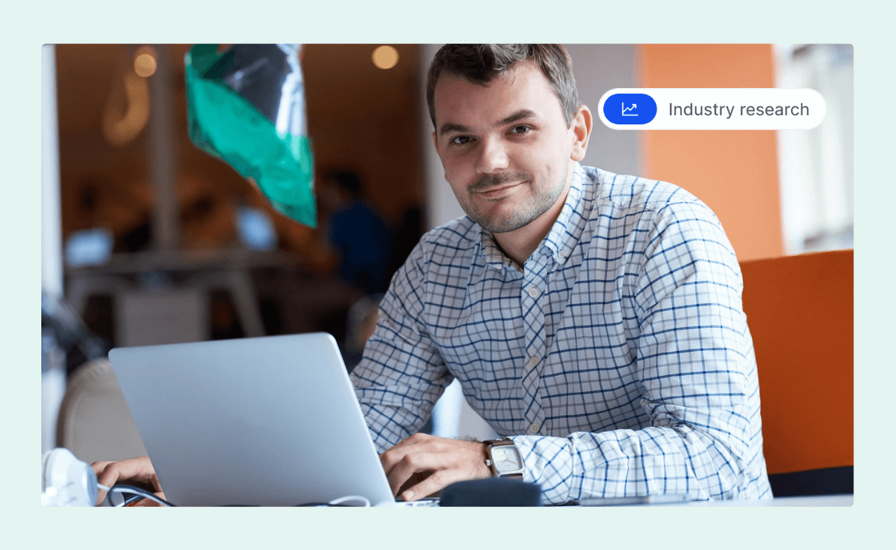 Man in a checkered shirt working on a laptop and smiling in a modern office.