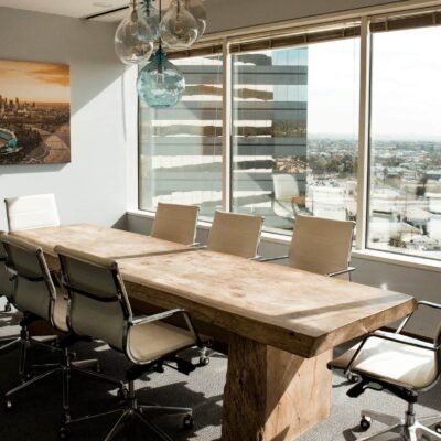 Empty meeting room with a large wooden table, white office chairs, and floor-to-ceiling windows overlooking a cityscape.