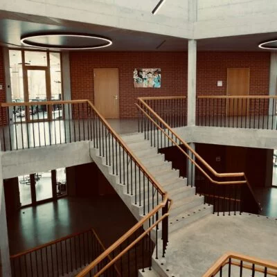 Interior of a modern building with exposed concrete, brick walls, and a central staircase surrounded by glass doors and classrooms.