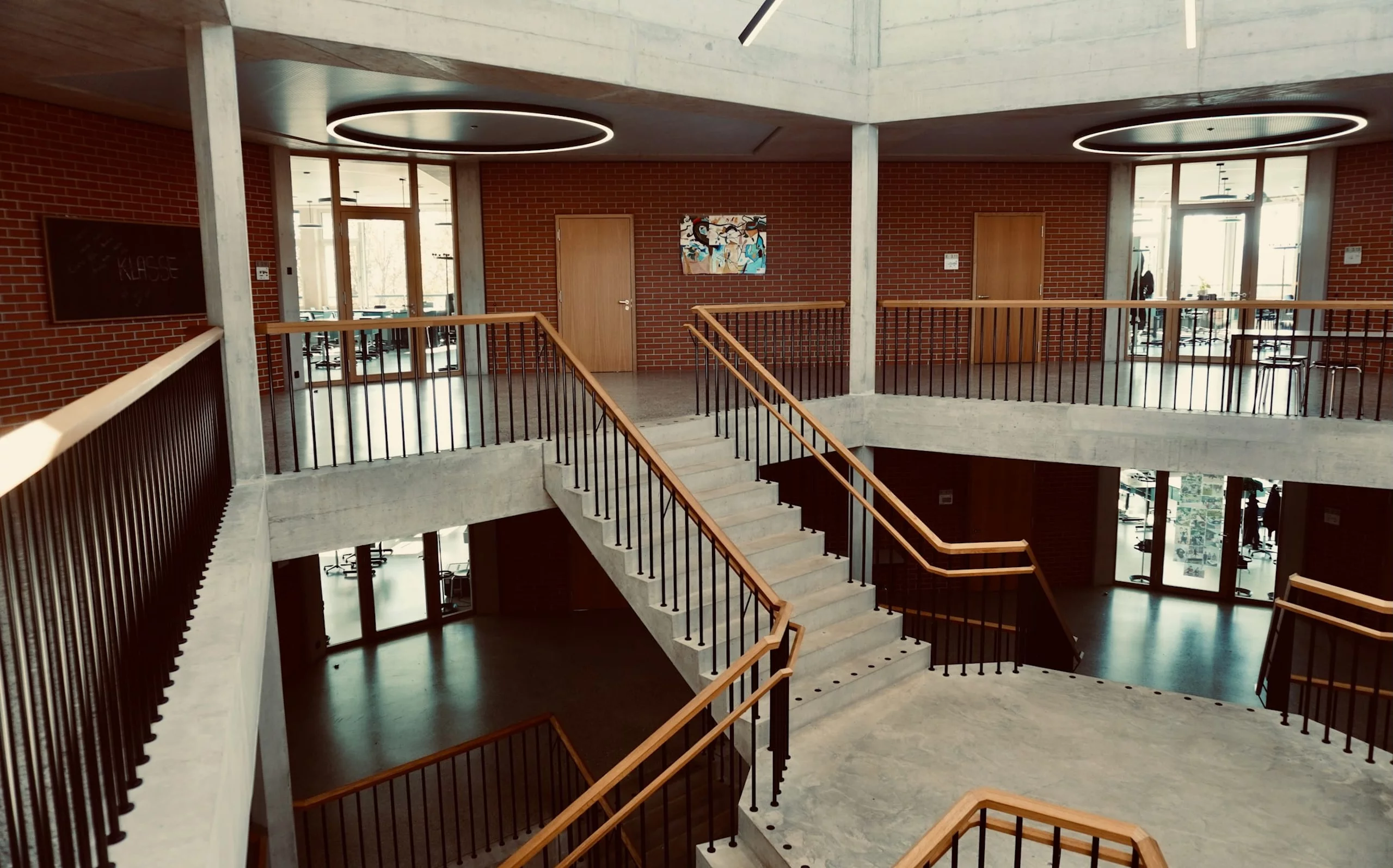 Interior of a modern building with exposed concrete, brick walls, and a central staircase surrounded by glass doors and classrooms.