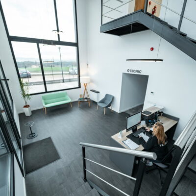 Reception area with a front desk, a staff member working, and large windows overlooking a parking lot.