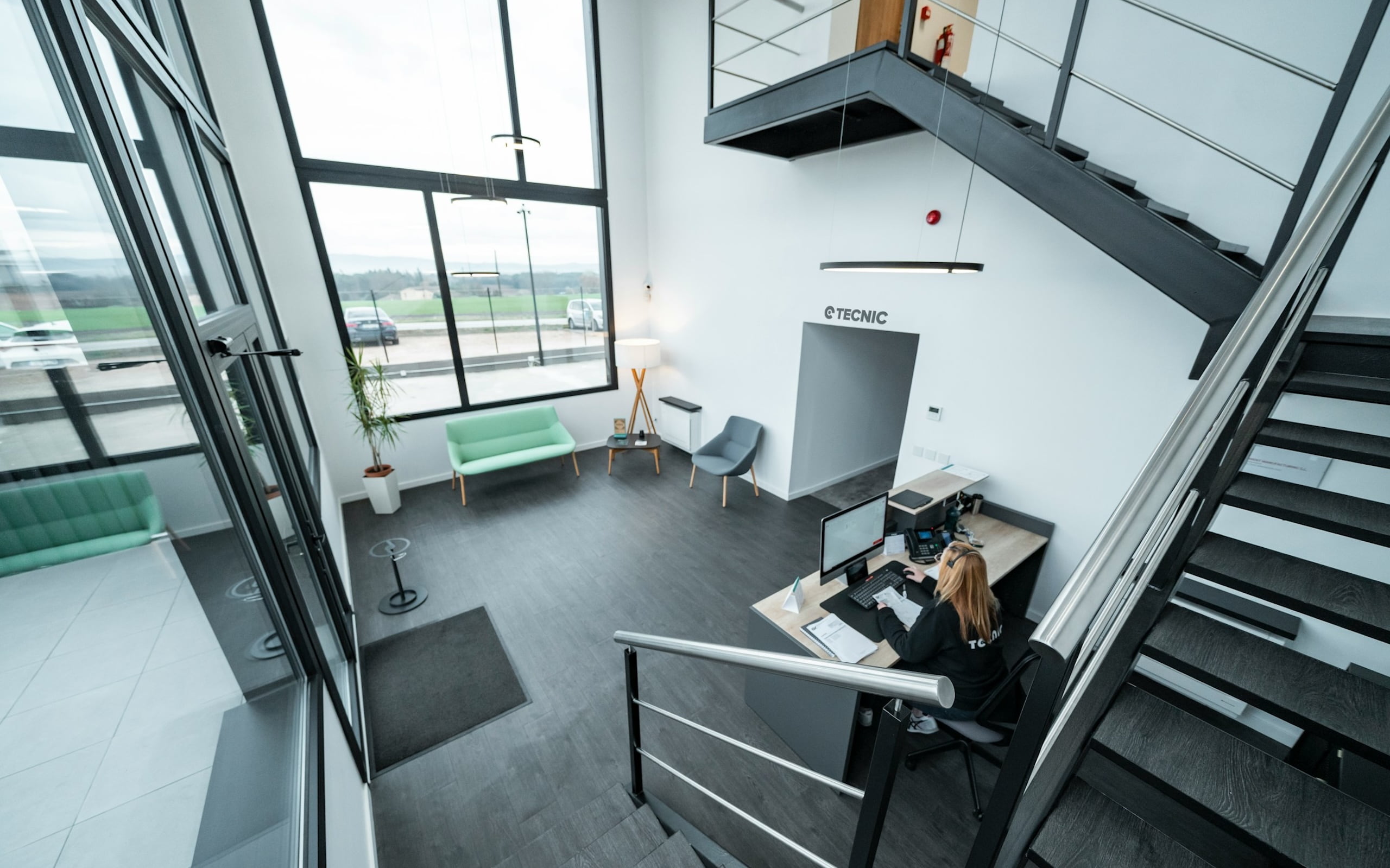 Reception area with a front desk, a staff member working, and large windows overlooking a parking lot.