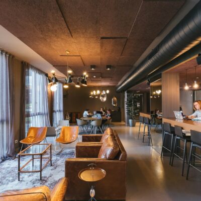 Modern coworking lounge with large windows, cozy seating, and people working at shared tables and bar-height desks.