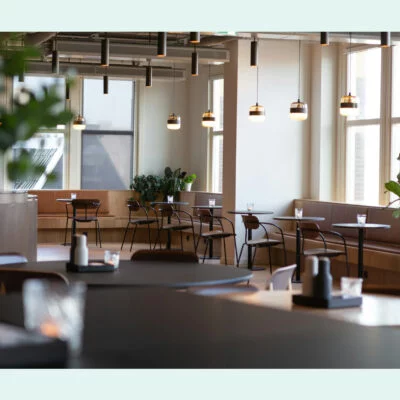 Modern office café with round tables, hanging lights, and potted plants by large windows.