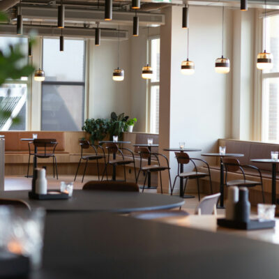 Moderna cafetería de oficina con mesas redondas, lámparas colgantes y plantas en macetas junto a grandes ventanales.