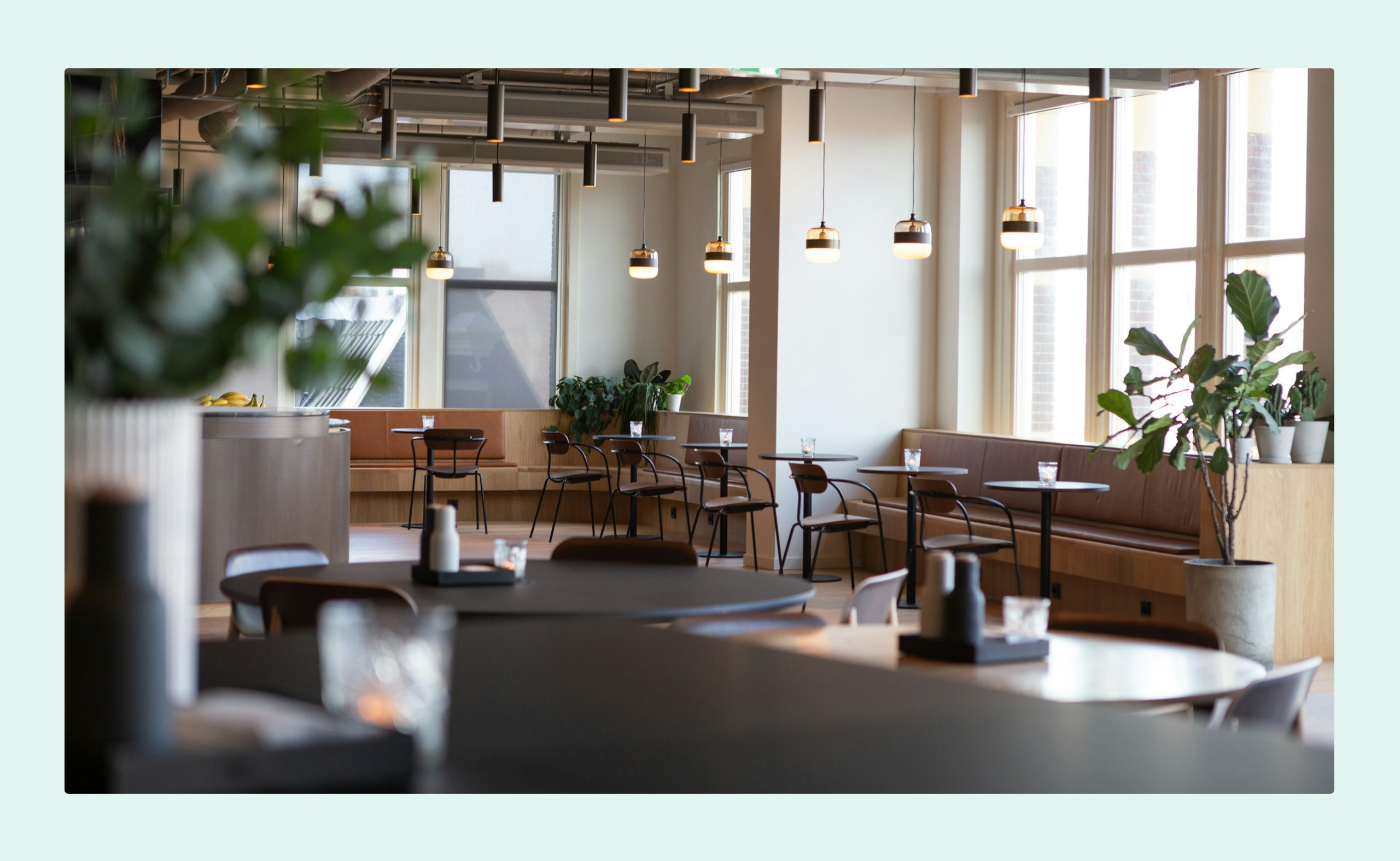 Modern office café with round tables, hanging lights, and potted plants by large windows.