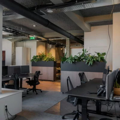 Modern open-plan office with black desks, computer monitors, and planters dividing the workspace.