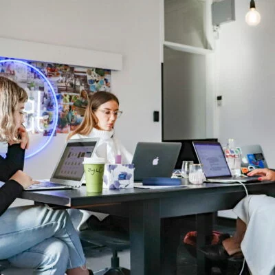 Three women sitting around a black table with laptops and drinks, working in a casual office.