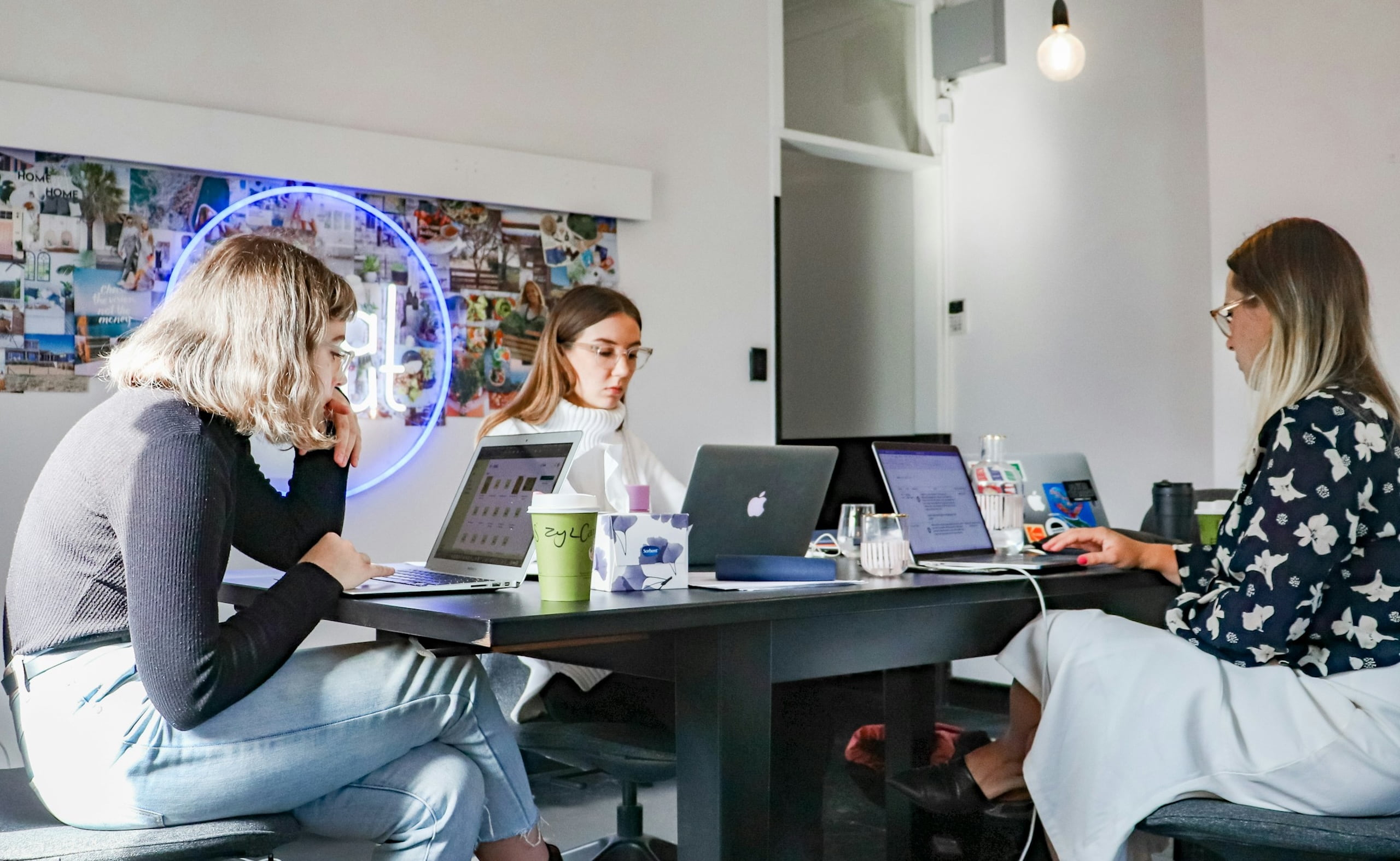 Three women sitting around a black table with laptops and drinks, working in a casual office.