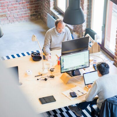 Two people working at a shared wooden desk in a bright, brick-walled office with striped rugs and large black pendant lamps.