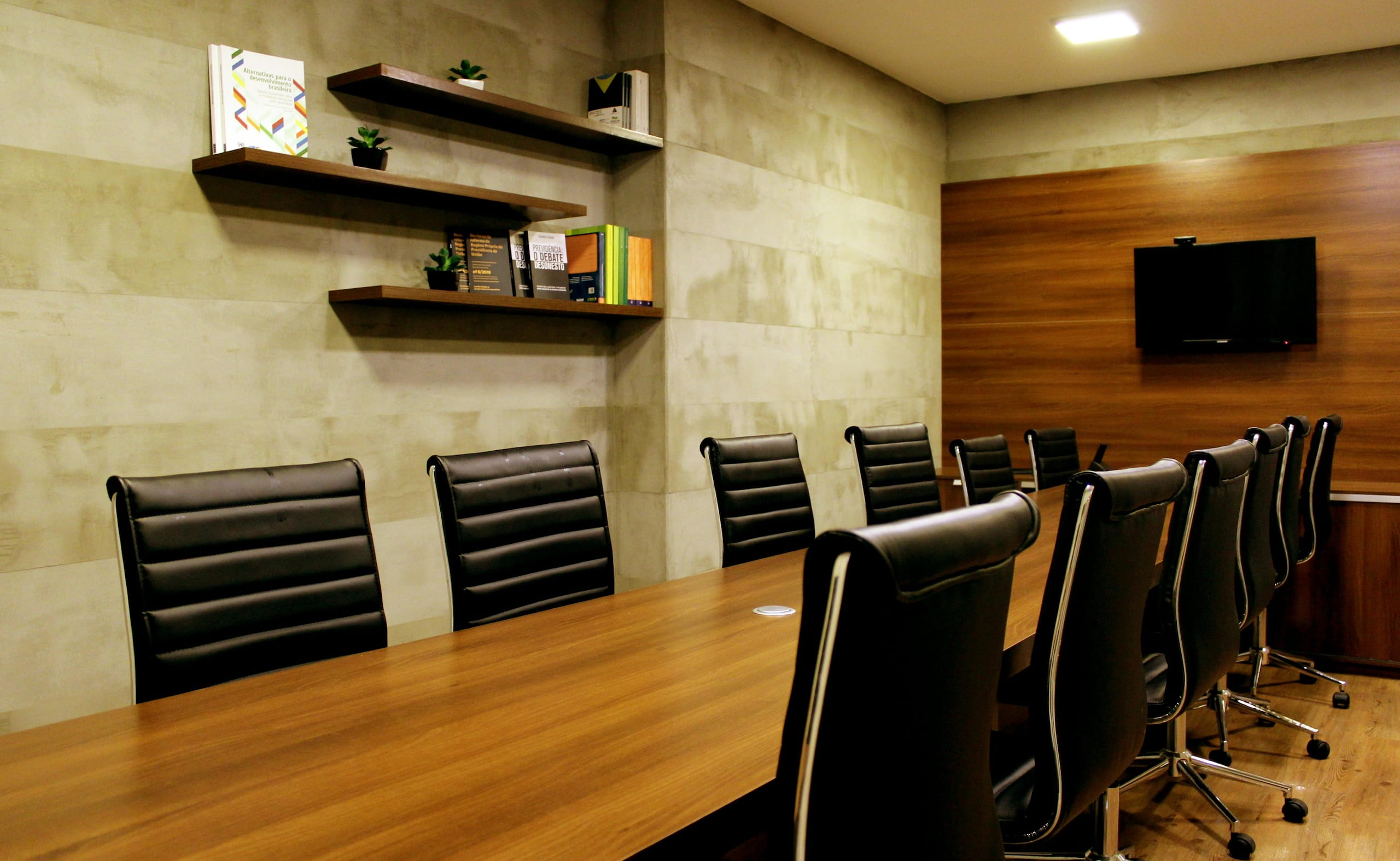 Modern conference room with a long wooden table, black chairs, bookshelves, and a wall-mounted TV.