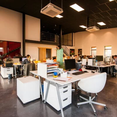 Open-plan modern office with white desks and chairs, where teams are working on computers in a bright space with large windows.