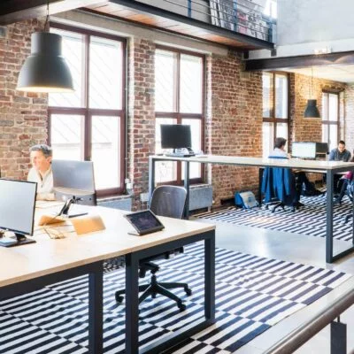 Bright loft-style office with brick walls, large windows, striped rugs, and several people working at shared desks with desktop monitors.