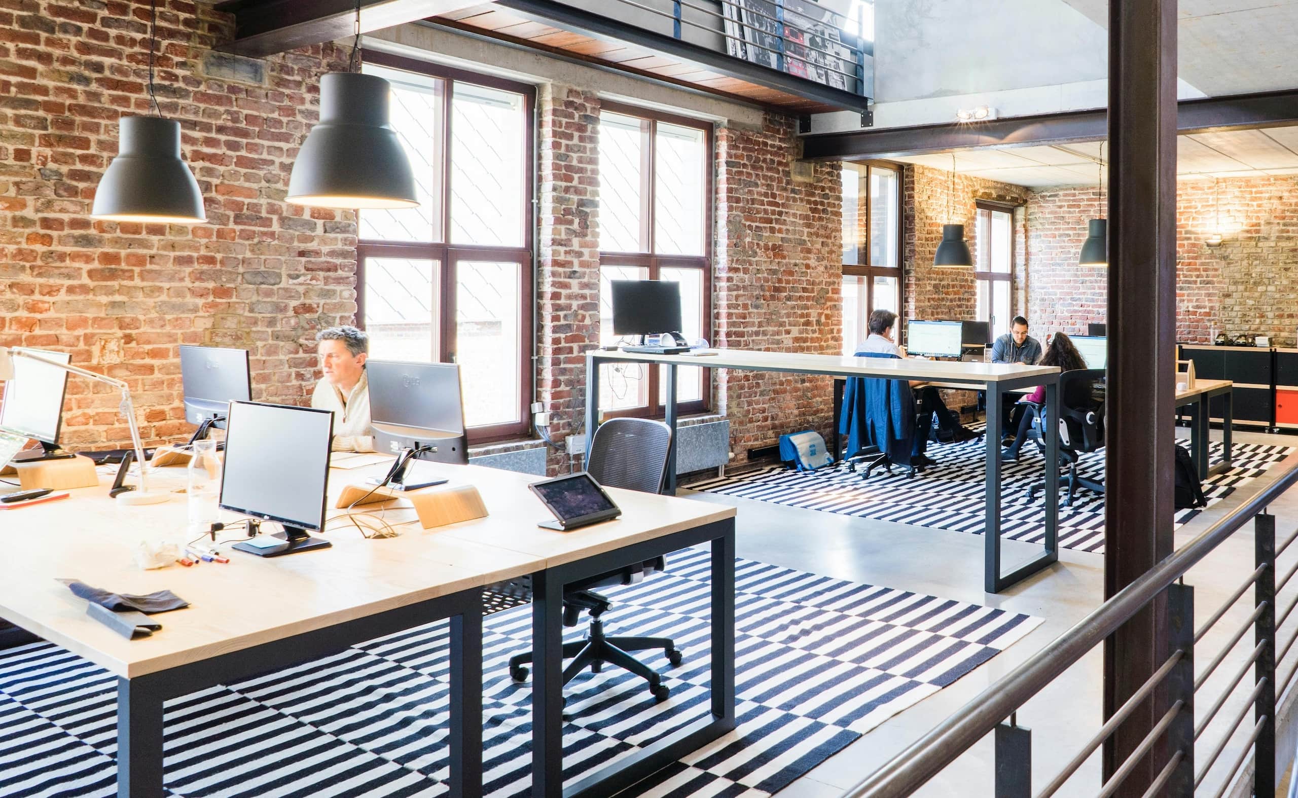 Bright loft-style office with brick walls, large windows, striped rugs, and several people working at shared desks with desktop monitors.