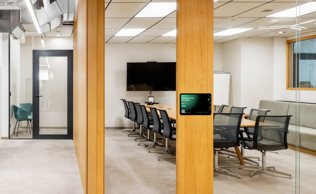 Modern meeting room with a large screen, glass walls, and a digital booking display panel by the door running the Archie app.