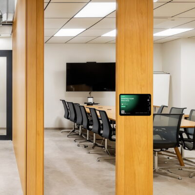 Modern meeting room with a large screen, glass walls, and a digital booking display panel by the door running the Archie app.