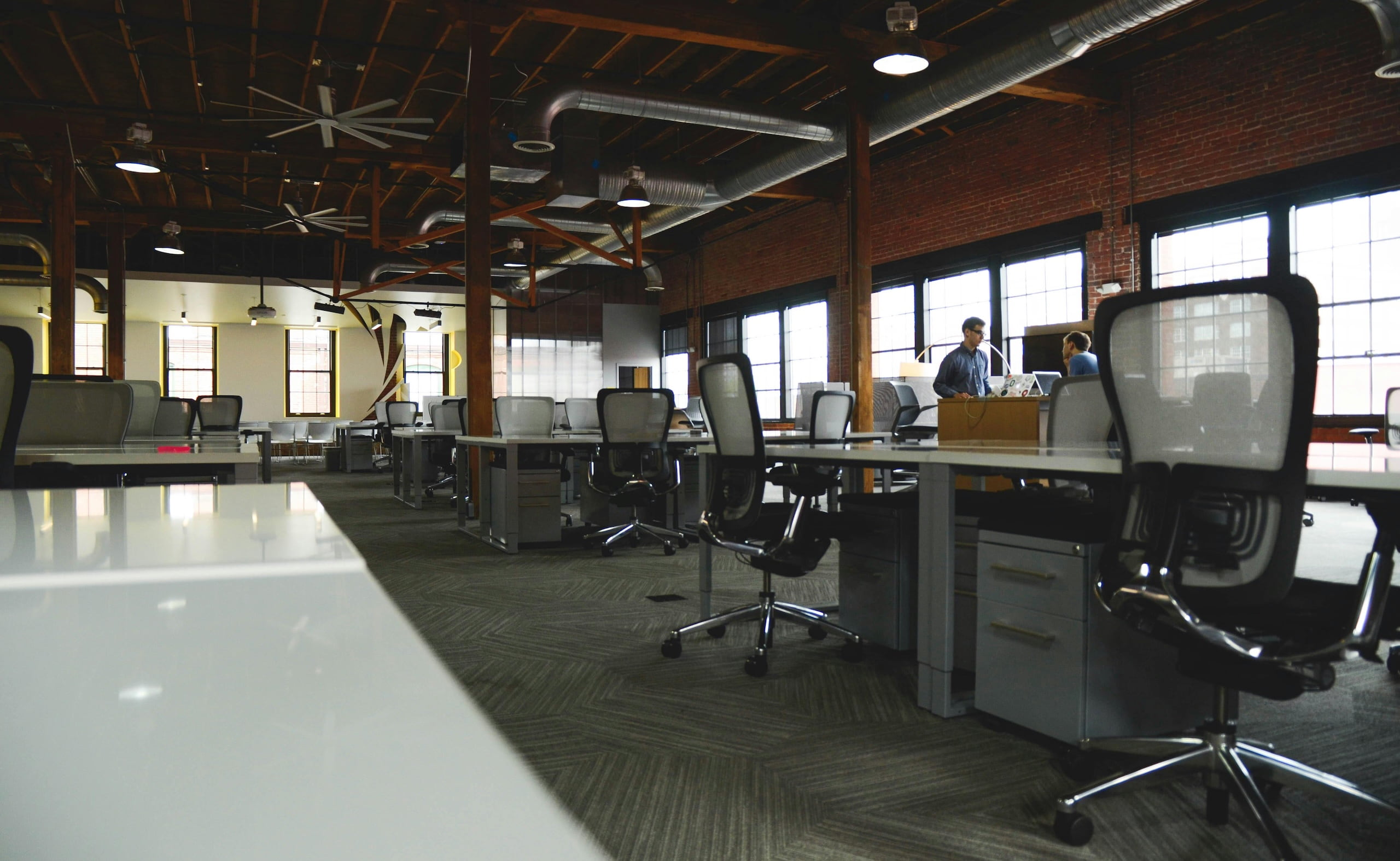 Spacious industrial-style office with exposed beams, large windows, and rows of empty desks and chairs, with two people talking in the background.