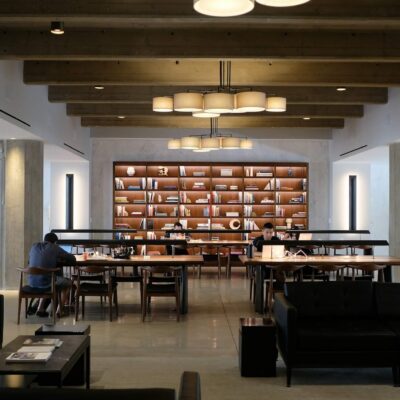 Open coworking lounge with long shared tables, bookshelves, and people working in a quiet space.