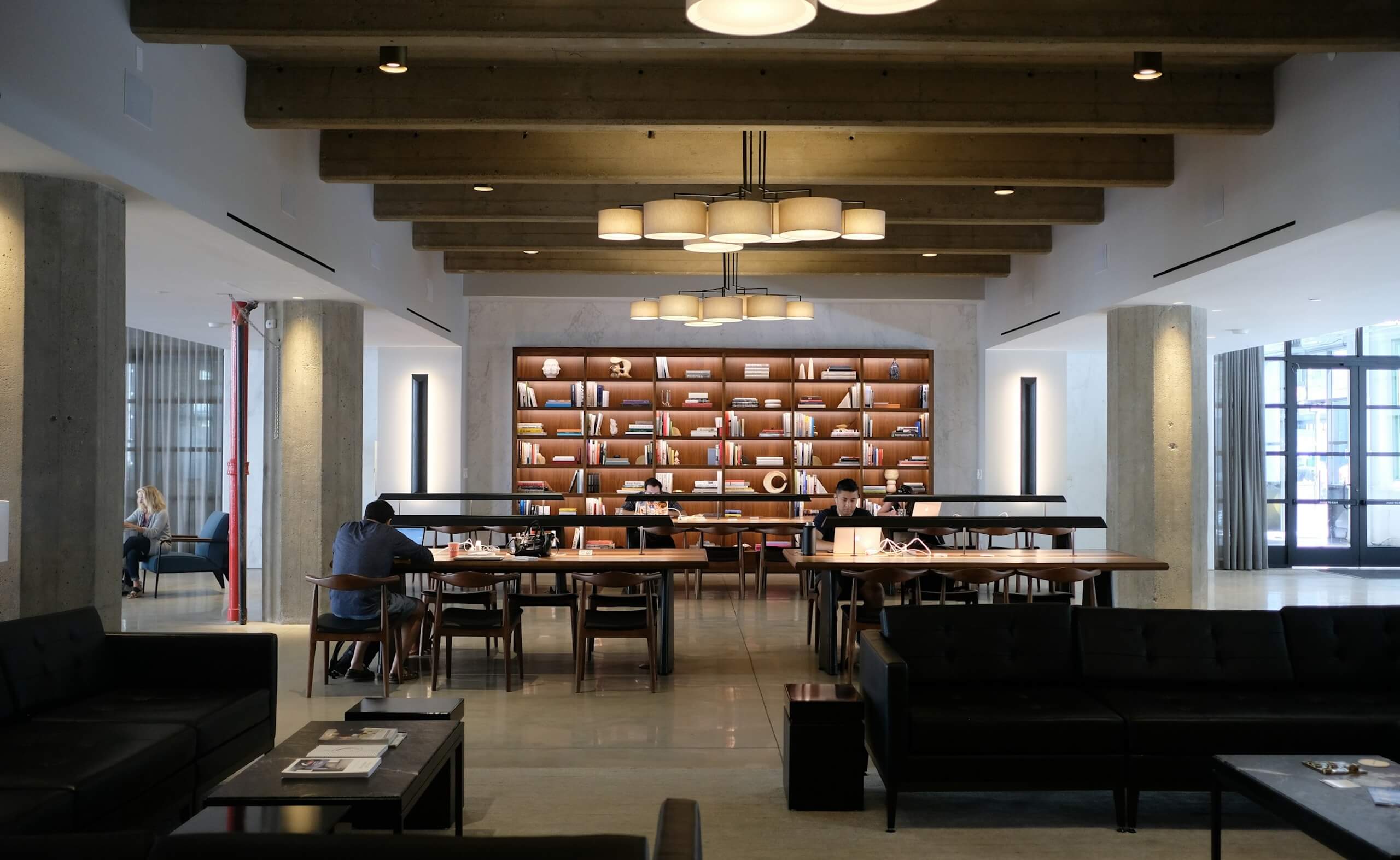 Open coworking lounge with long shared tables, bookshelves, and people working in a quiet space.